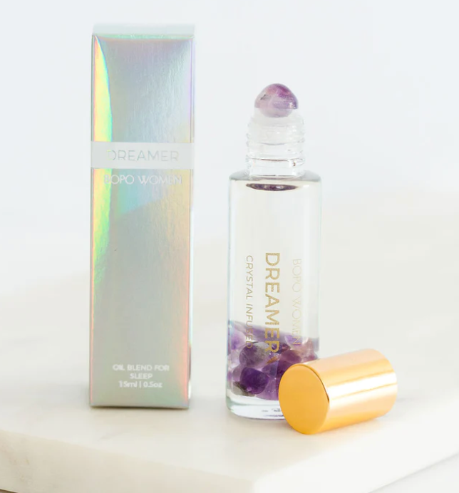 A clear glass roll-on bottle labeled 'DREAMER' with purple crystal pieces inside and a matching iridescent box beside it, plus a gold cap resting in front, set on a white surface against a white background.