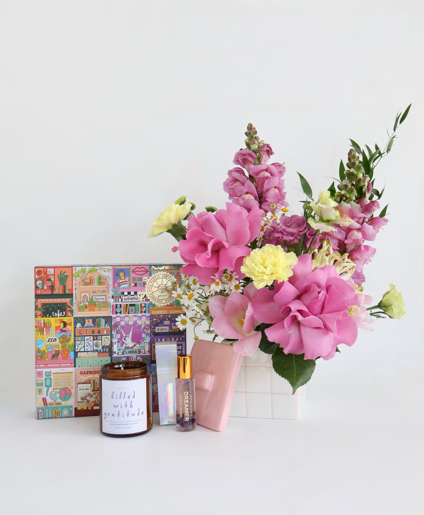 A styled product arrangement on a white surface against a white background, featuring a colourful pink and yellow flower arrangement in a white grid-patterned ceramic that comes with a lid for a keepsake trinket box,  a small handmade soy wax candle in amber jar, and a colorful illustrated Astrology 500 piece puzzle.